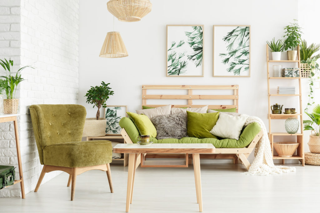 Green armchair next to wooden table in floral living room with lamps and pillows on sofa against white wall with leaves posters